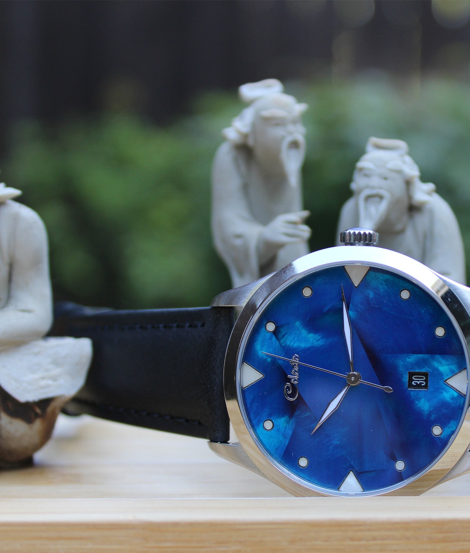Wristwatch with cobalt blue face and black strap on a wooden surface with stone statues in the background