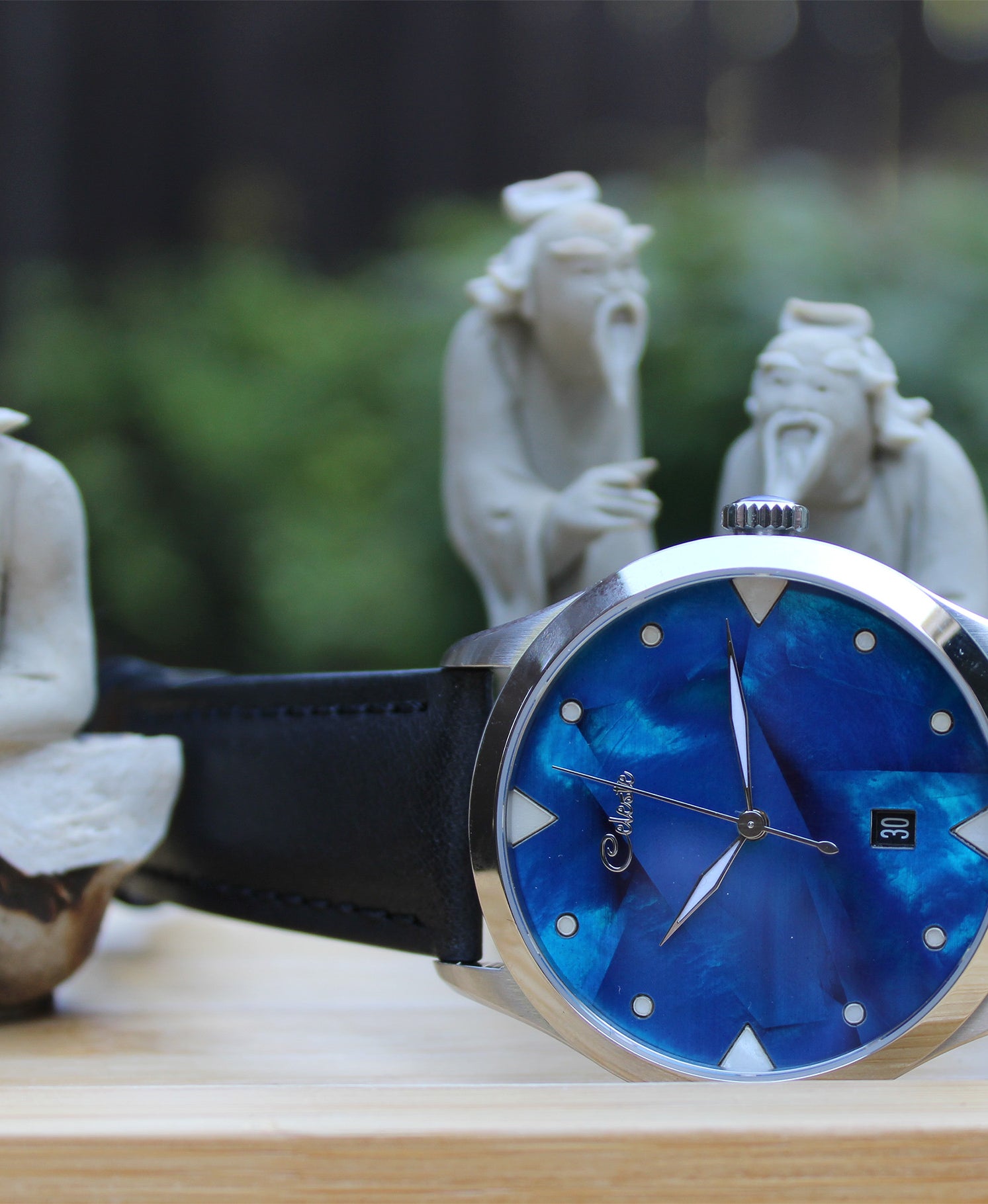 Wristwatch with cobalt blue face and black strap on a wooden surface with stone statues in the background