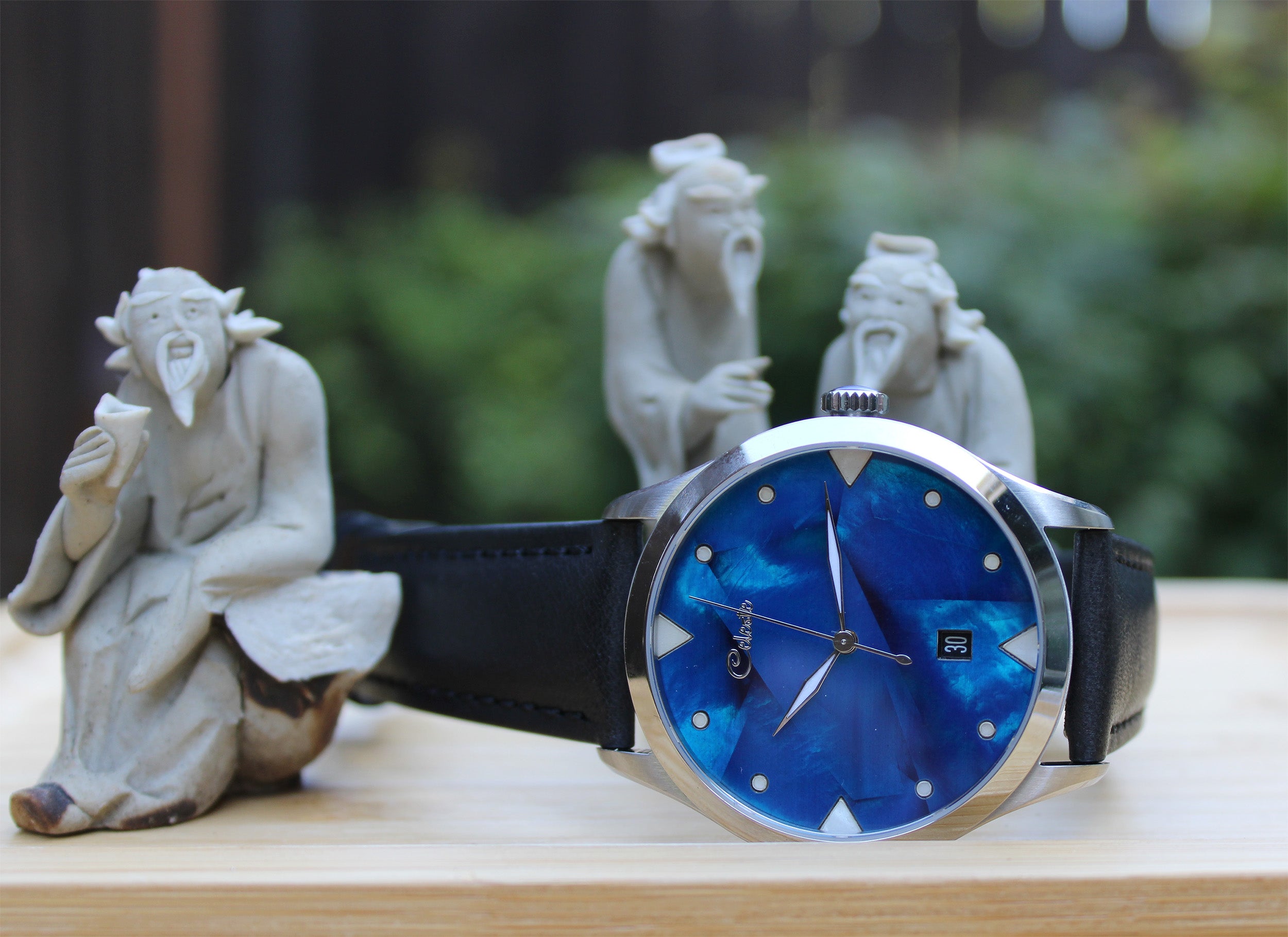 Wristwatch with cobalt blue face and black strap on a wooden surface with stone statues in the background